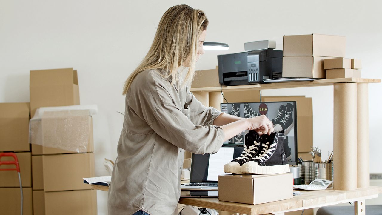 Woman holding a pair of black shoes above an open shipping box while packing an online store order